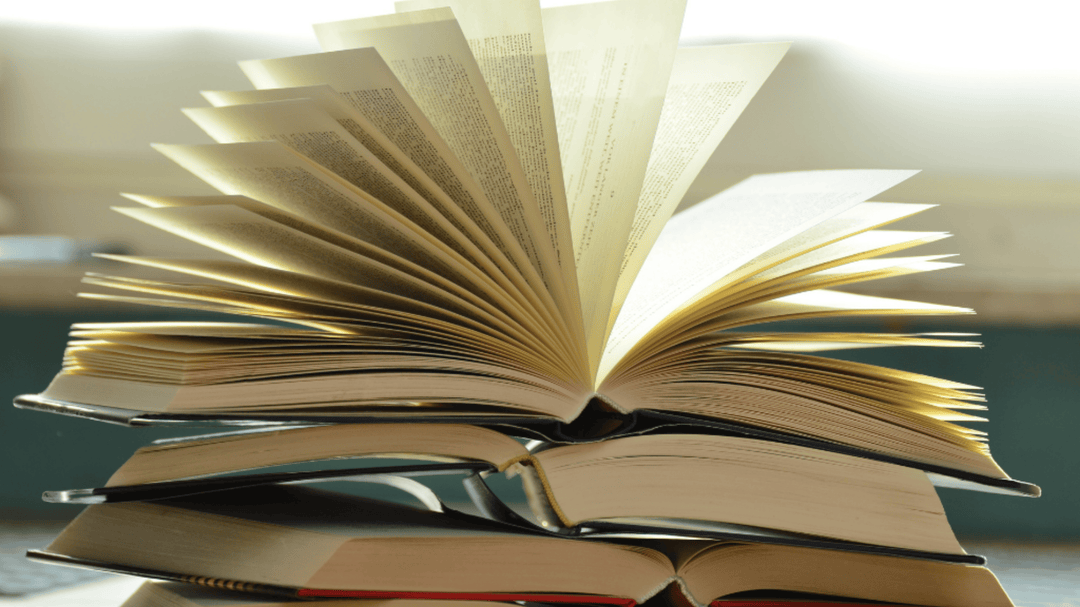 A close-up shot of a stack of open books with one book in the middle fanned out dramatically, pages fluttering upward as if caught by a breeze, with soft natural light in the background.