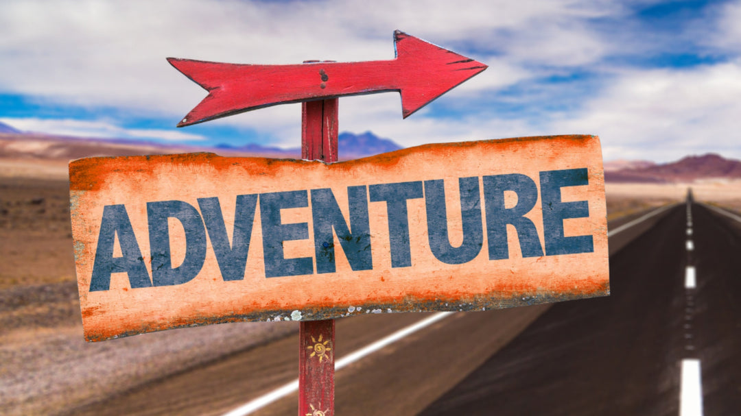 A weathered wooden sign with the word “ADVENTURE” in bold blue letters points right along a desert highway under a bright blue sky.