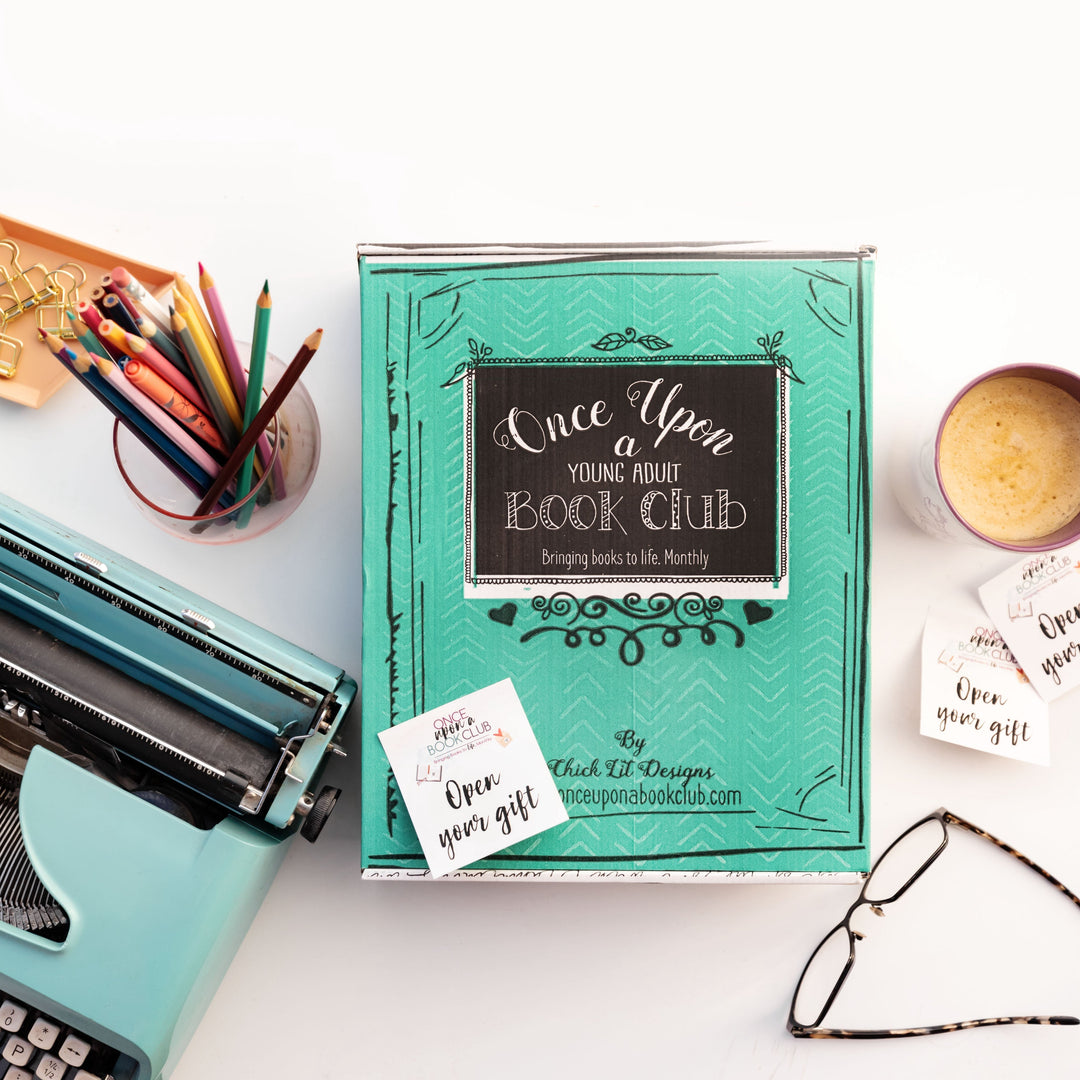 A teal Once Upon a Book Club box sits on a desk with a typewriter, colorful pencils, glasses, and a cup of coffee nearby.