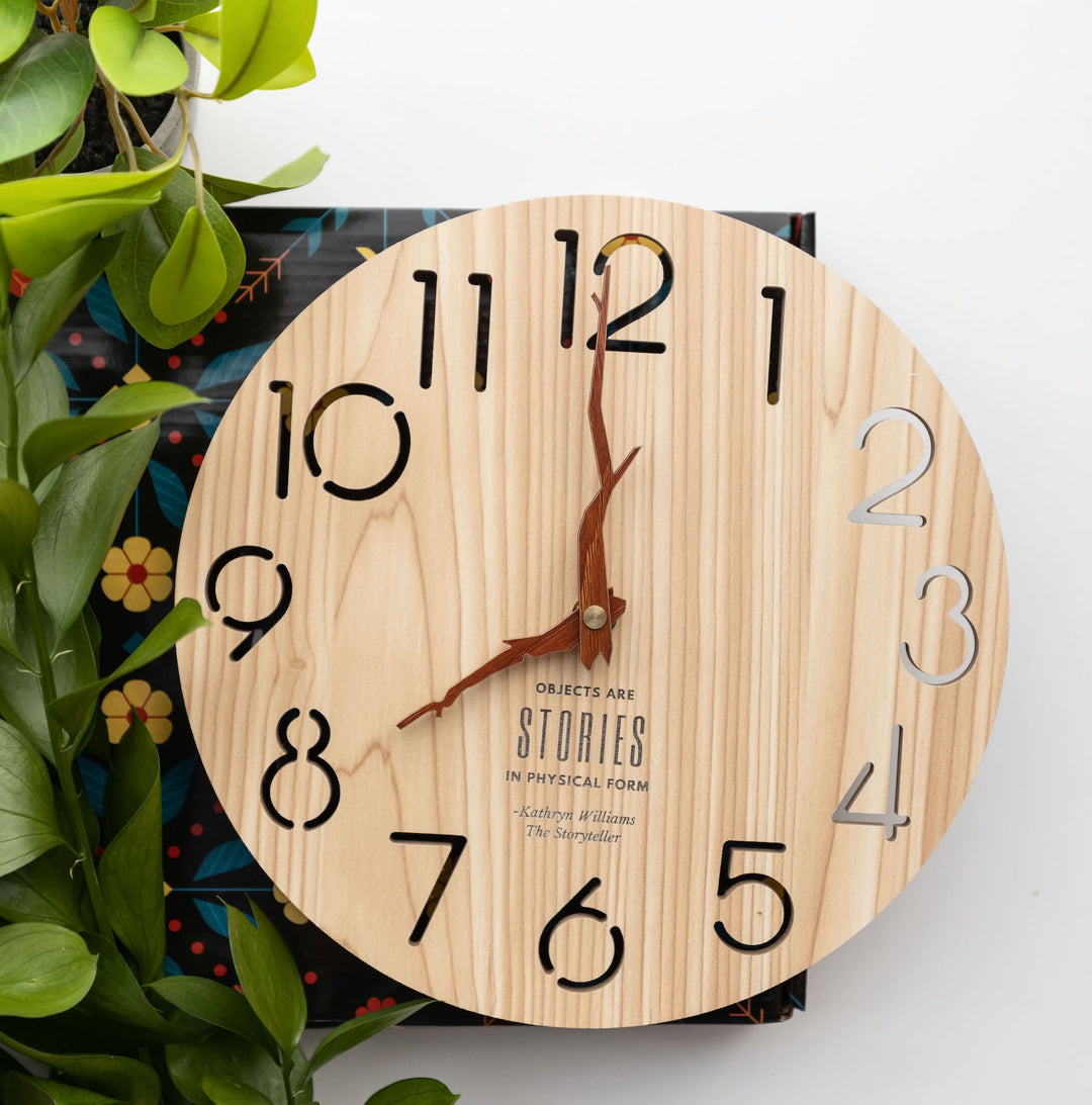 A round wooden clock with carved numbers and rustic twig-style clock hands, accompanied by leafy greenery on a decorative background.