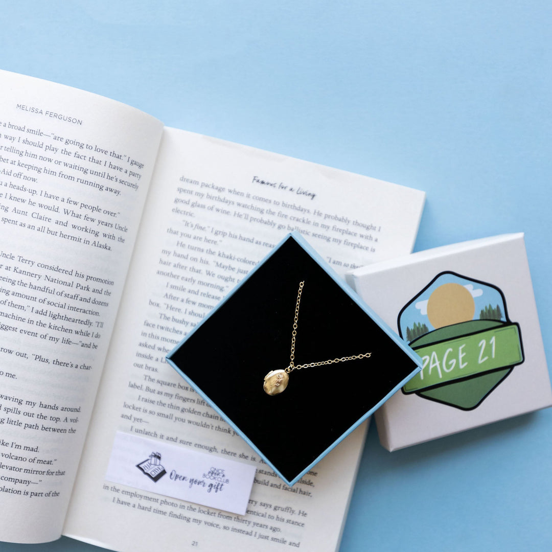 An open book lies next to a delicate gold necklace displayed in a box, accompanied by a matching gift box labeled "Page 21."