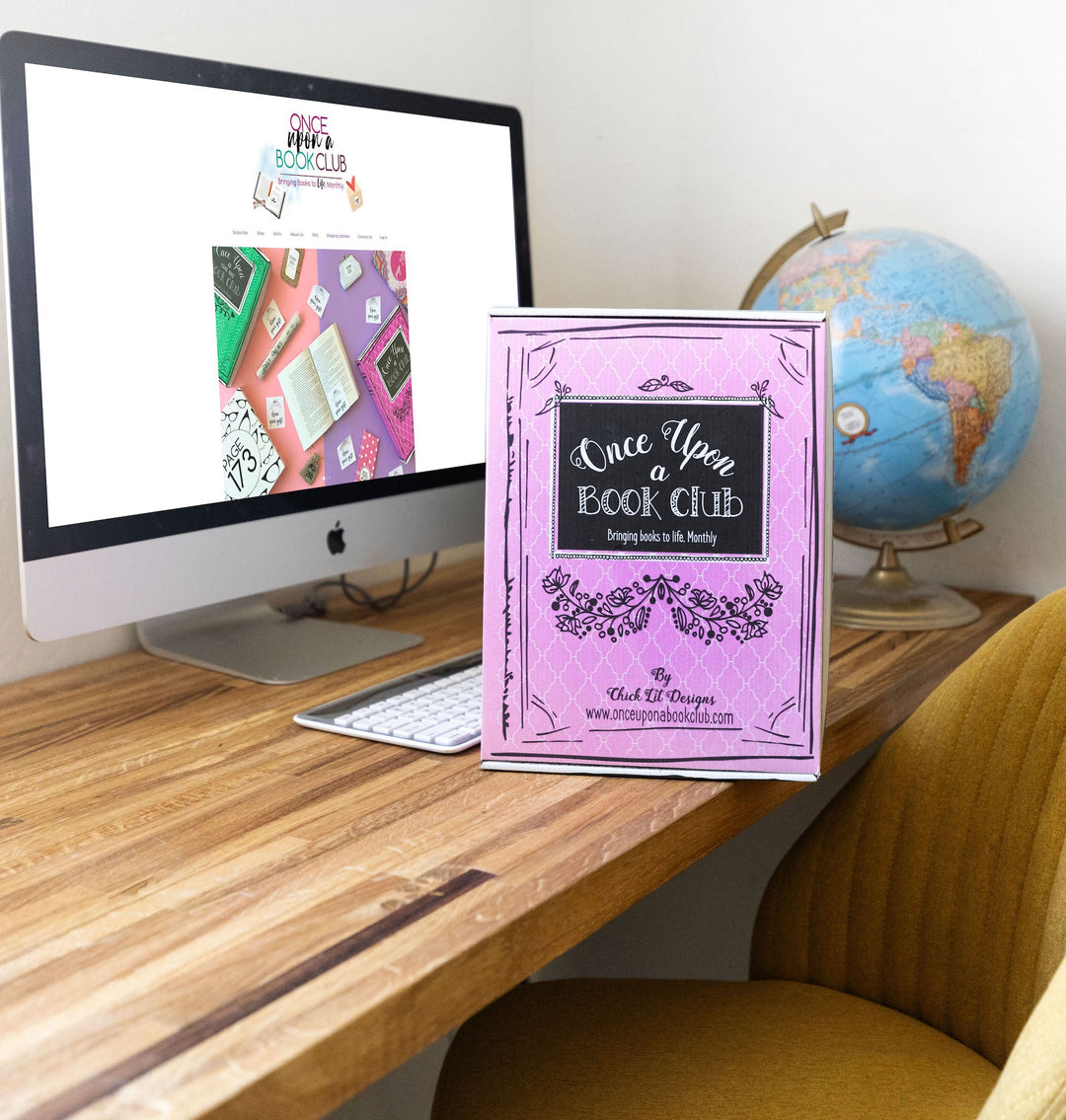 A Once Upon a Book Club box displayed on a wooden desk with a computer screen showing the website and a globe in the background.