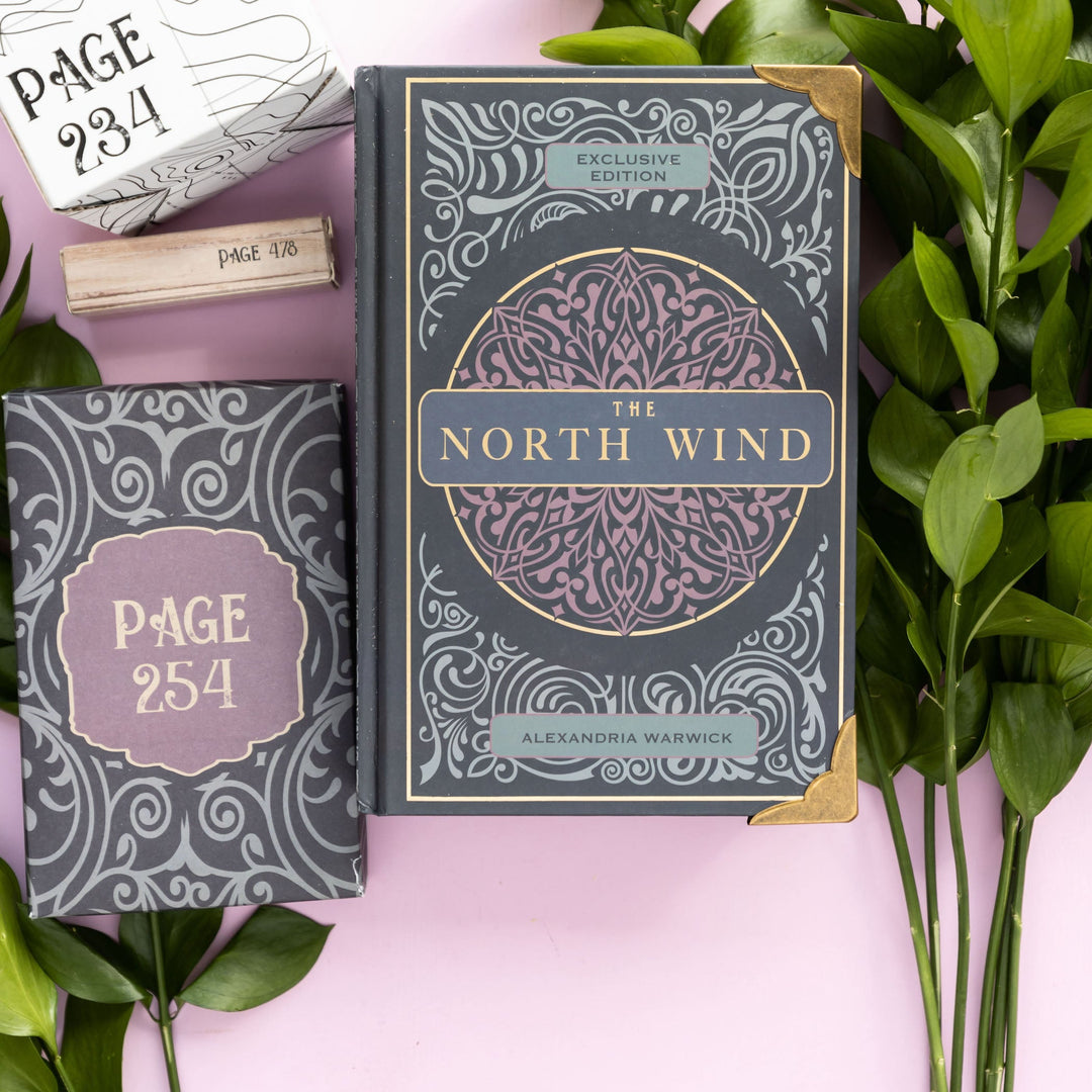 Deluxe edition of The North Wind by Alexandria Warwick with gold detailing, book accessories, and green leaves on a pink background.