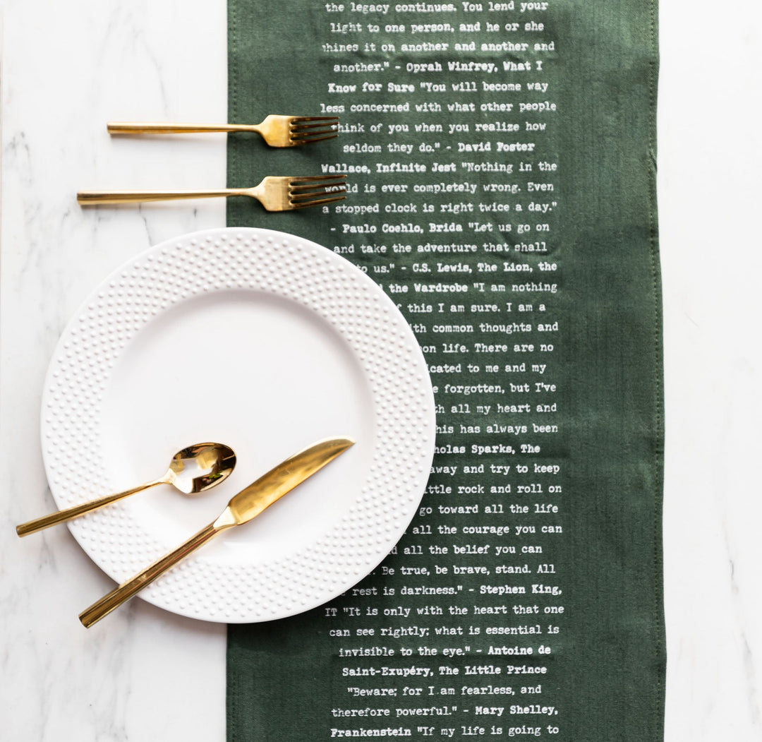 A table setting featuring a white textured plate, golden utensils, and a green table runner printed with famous literary quotes.
