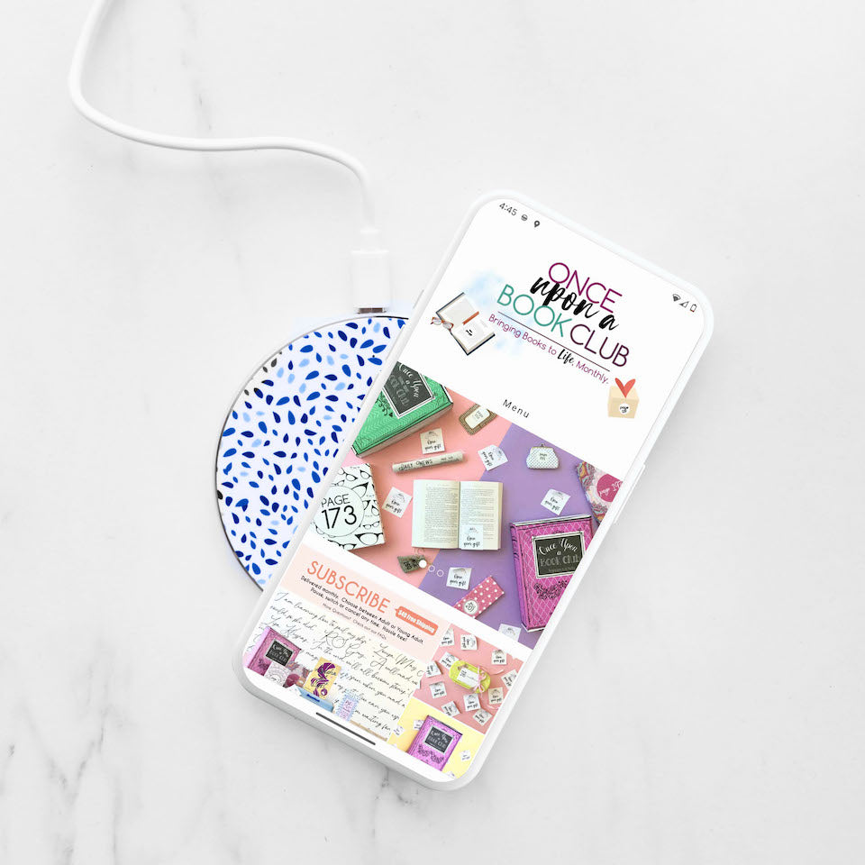 A smartphone displaying the Once Upon a Book Club website rests on a wireless charger with a blue and white patterned surface.