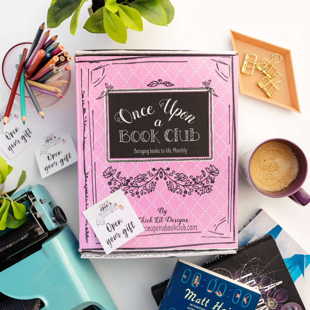 Pink Once Upon a Book Club box surrounded by colorful pencils, books, and a coffee mug, showcasing a creative reading subscription.