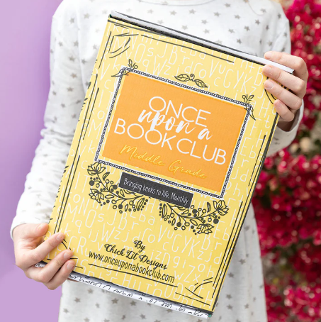 A yellow "Once Upon a Book Club" Middle Grade box with a bold orange label is held by a person wearing a white and gray polka-dot outfit.