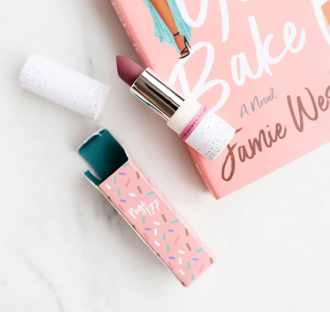 A pink-themed setup featuring a novel by Jamie Wesley, a matte lipstick with sprinkles design packaging, and a beauty-inspired aesthetic.