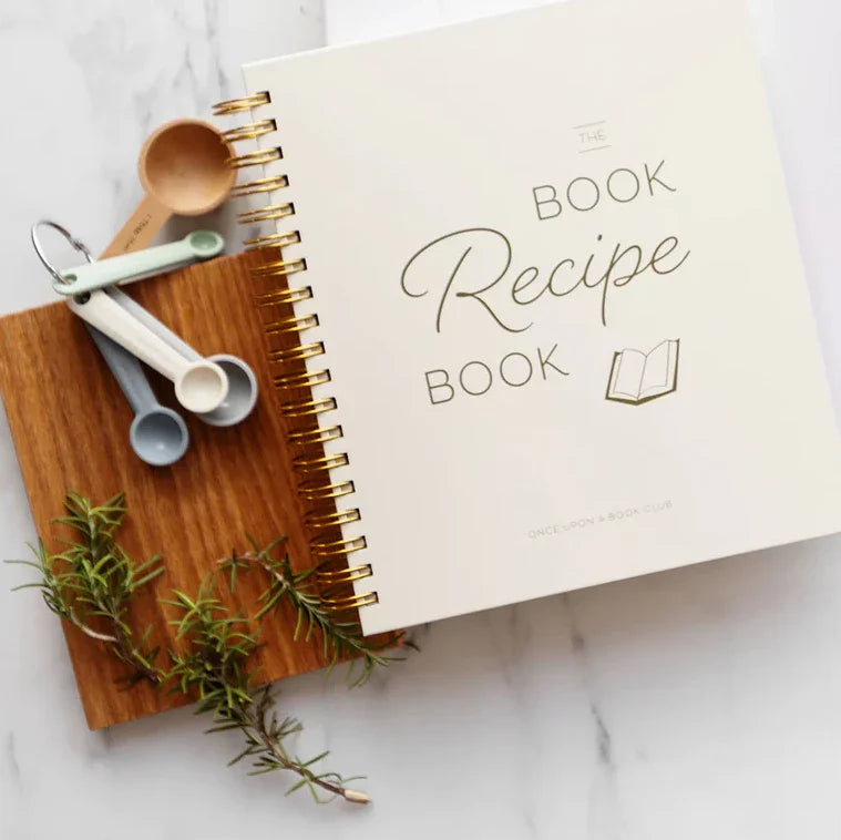 A spiral-bound recipe book titled "The Book Recipe Book" lies on a wooden cutting board surrounded by measuring spoons and sprigs of rosemary.