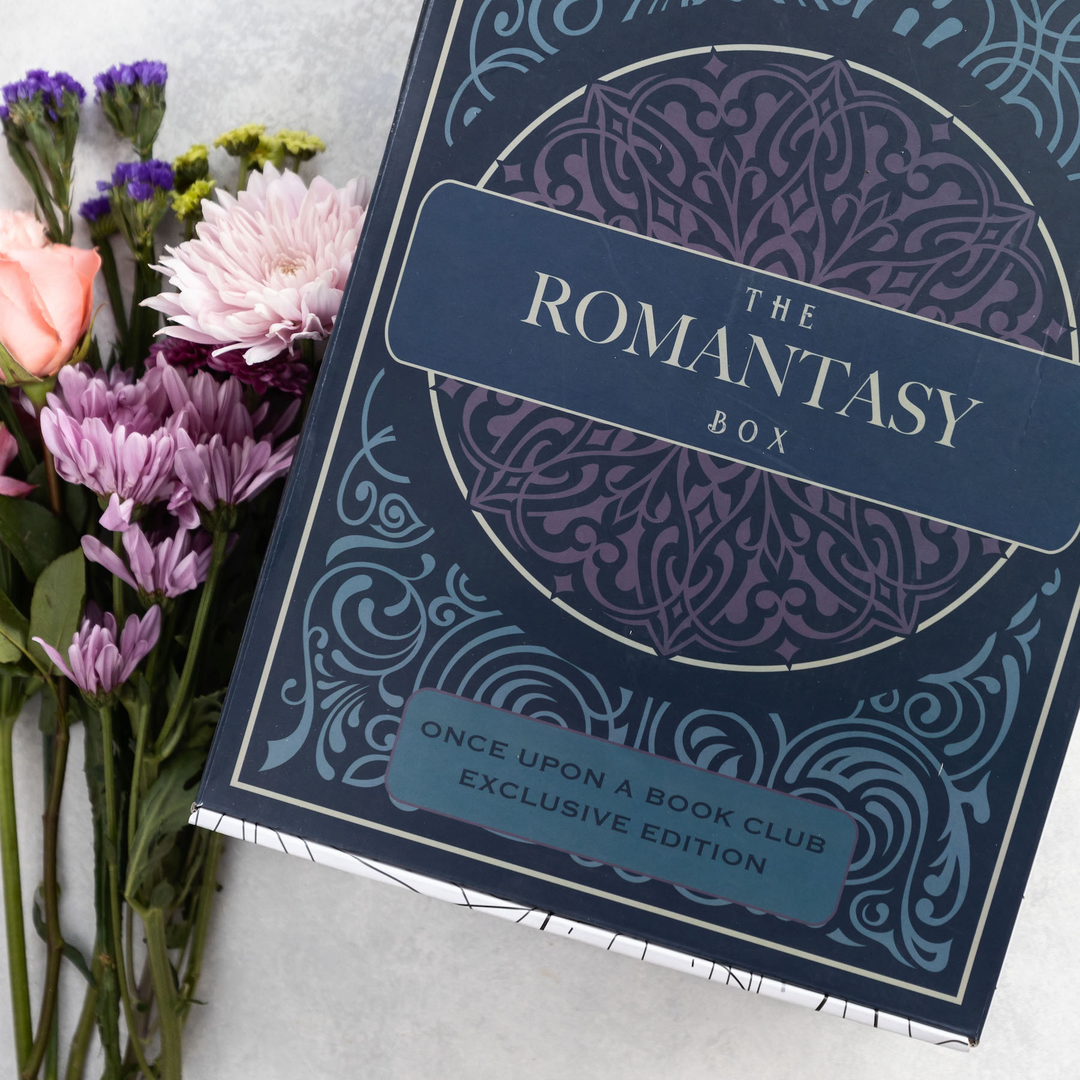 A navy blue book box labeled "The Romantasy Box" featuring intricate patterns is placed beside a bouquet of colorful flowers on a light surface.