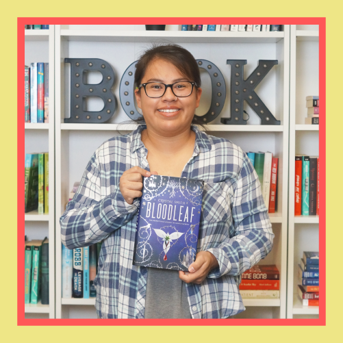 a dark-skinned woman with dark hair wearing glasses and a flannel stands in front of a bookshelf, smiling and holding a book
