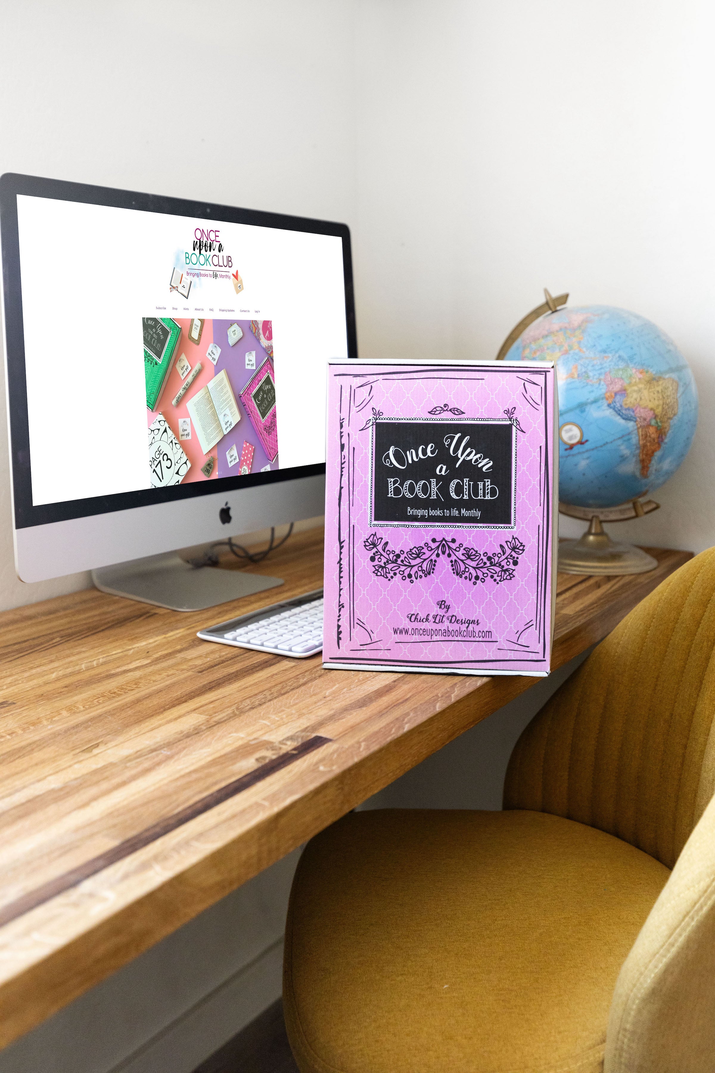 A pink Once Upon a Book Club box sits on a computer desk in front of a computer monitor. The monitor is displaying the Once Upon a Book Club website home page.