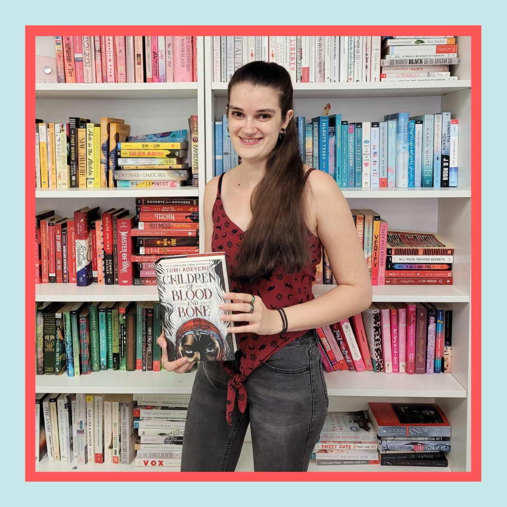 a white woman with dark brown hair wearing a red tank-top and black jeans stands in front of a bookshelf, smiling and holding a book