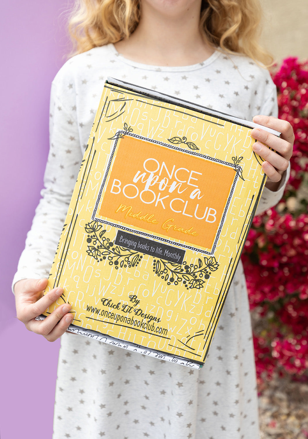 a white child wearing a white dress with gold stars holds a yellow Once Upon a Book Club box in front of her