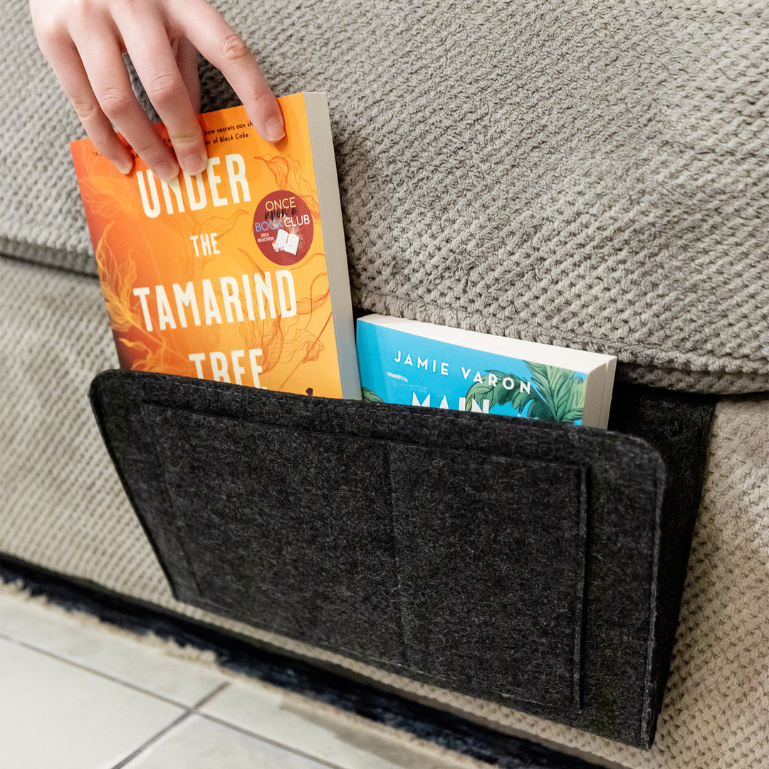 A fabric organizer sticks out from under a couch cushion. Inside are two books. A white hand pulls one of the books from the organizer.