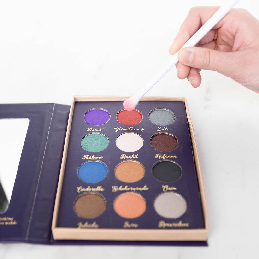A purple makeup pallet displaying a collection of 12 colors on a white background. A white hand holds a white makeup brush over the pallet.