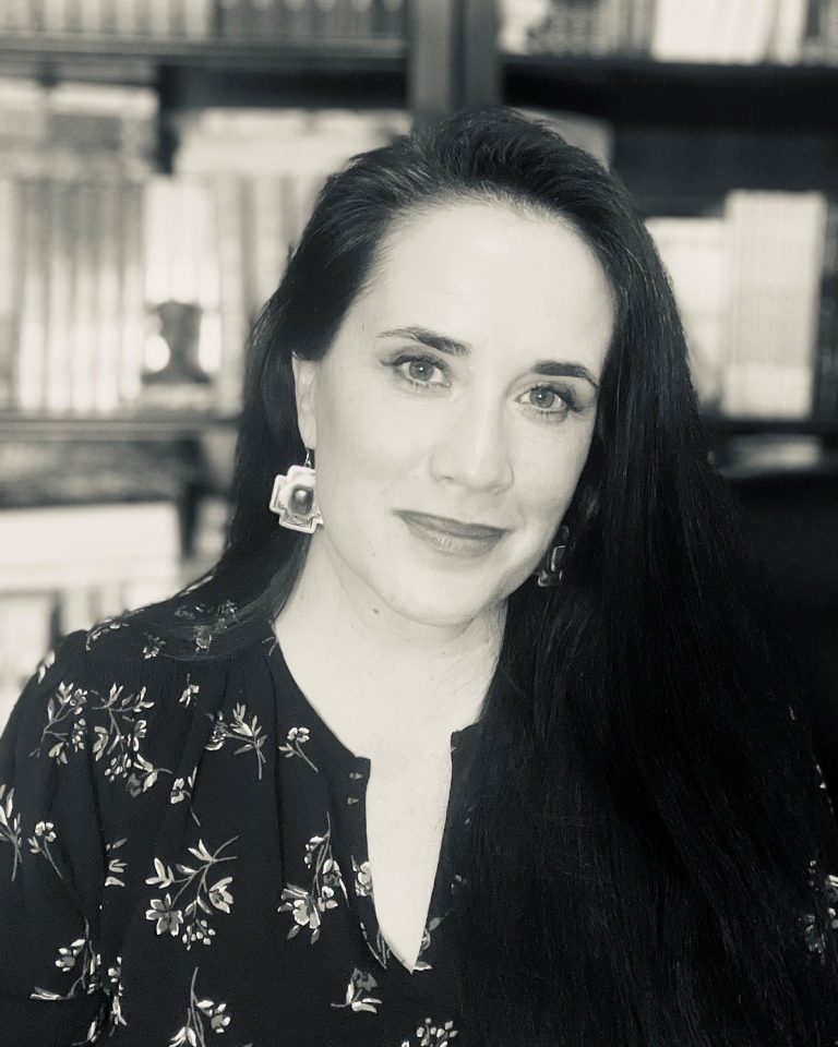 Author Amy Harmon is a young white woman with long dark hair. She is photographed in black and white and is wearing a dark, floral top. She is smiling at the camera.