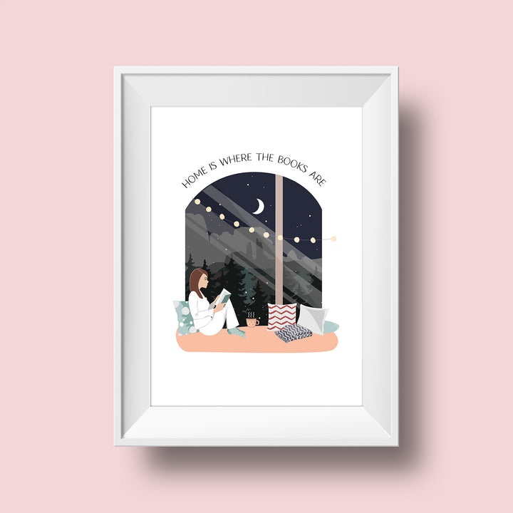 An art print hangs on a pink wall inside a white matted frame. The print features a white background with an illustration of a large window showing a nighttime scene including cliffs and trees in the distance. Over the window is the quote "Home is Where the Books Are" in black font. A crescent moon is visible in the night sky. In the window sits a brunette girl in cozy attire reading with a steaming mug next to her and pillows in various patterns around her.