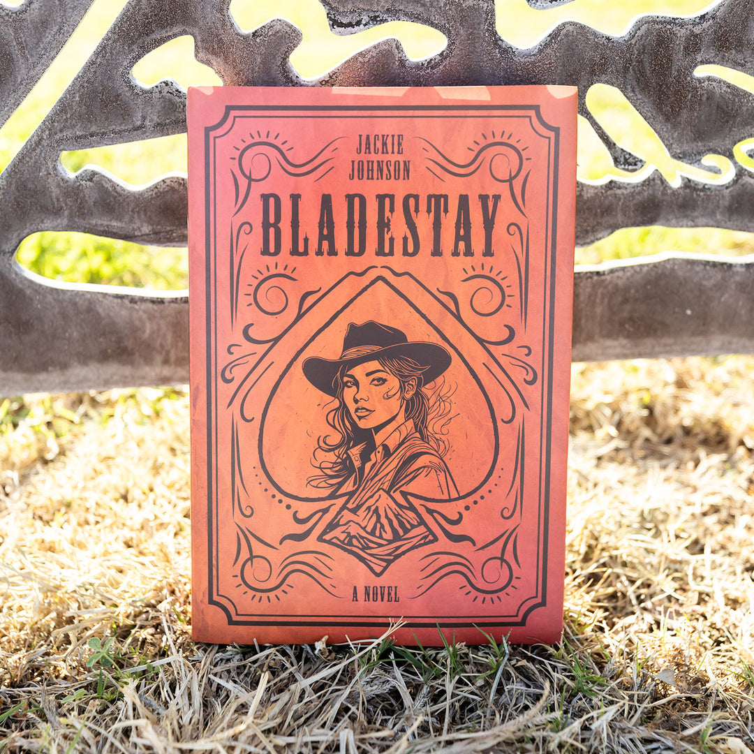 A hardcover, exclusive edition of Bladestay by Jackie Johnson outdoors on grass with a metal fence behind it.