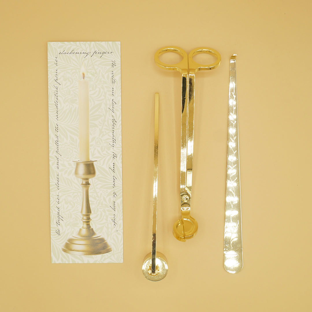 A gold candle care set - a wick trimmer, candle snuffer, and wick dipper next to a box with an image of a candlestick on it