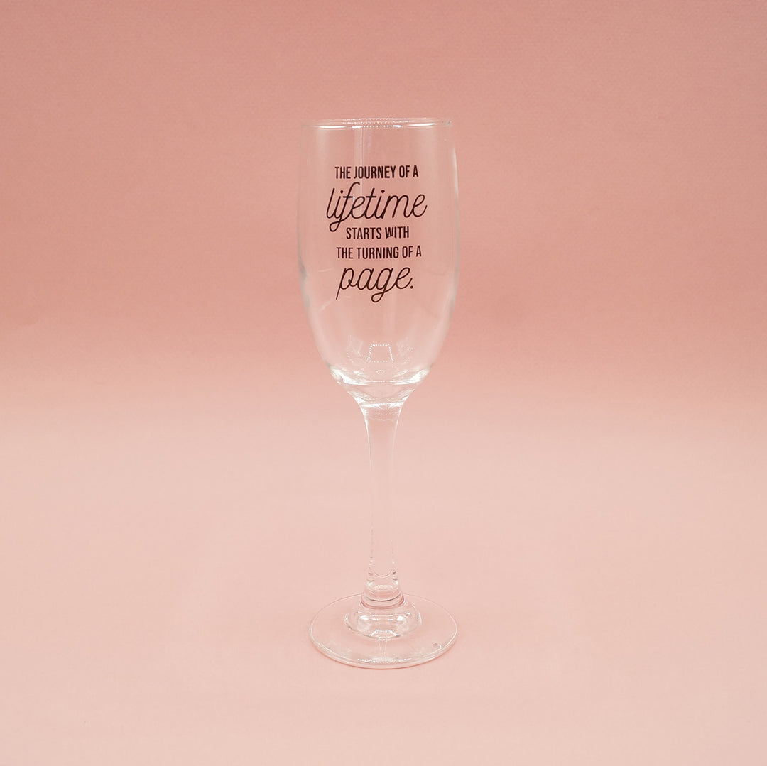 A single champagne flute with the words "The journey of a lifetime starts with the turning of a page" written on the glass sits on a pink background.