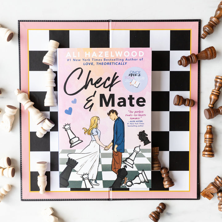 A paperback copy of Check & Mate by Ali Hazelwood sits in the middle of a pink bordered chess set surrounded by chess pieces.