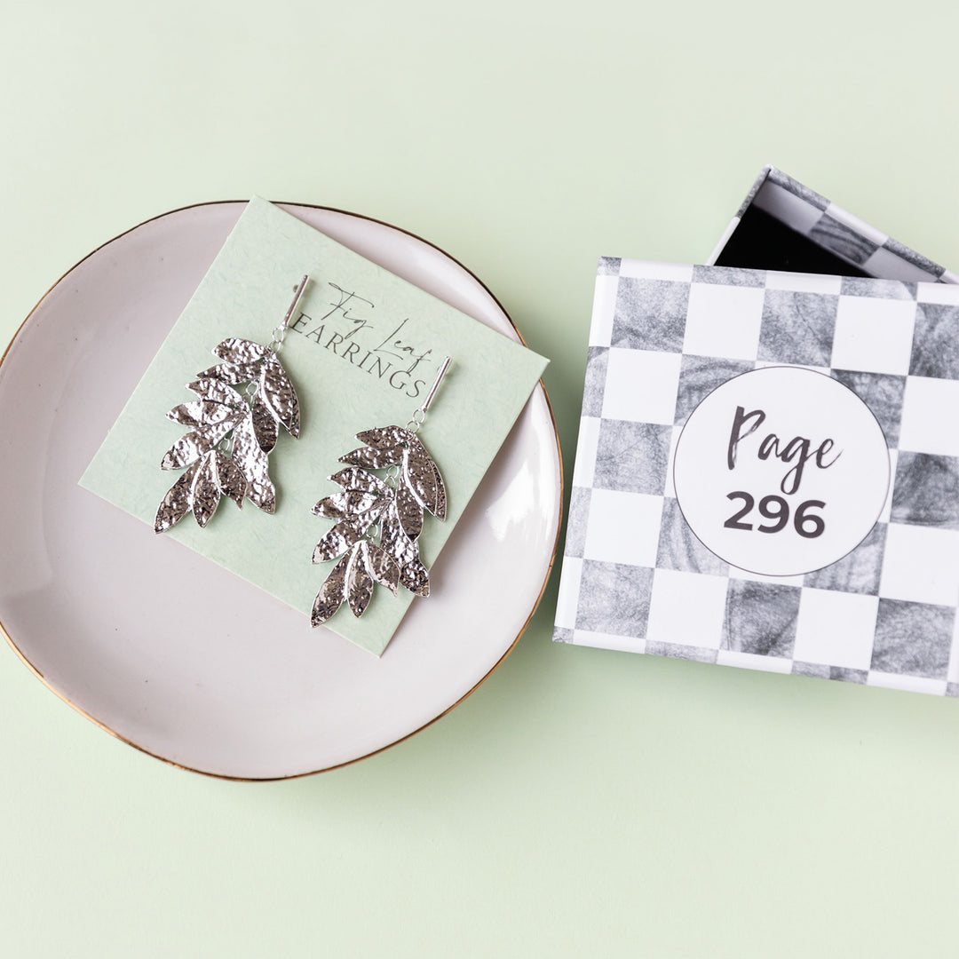 A pair of silver fig leaf dangle earrings sit on a green earring card inside a white trinket dish. An open box reading Page 296 sits next to the earrings.
