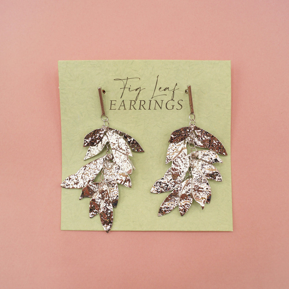 A close up of the silver dangle fig leaf earrings on a green earring card. On a pink background.