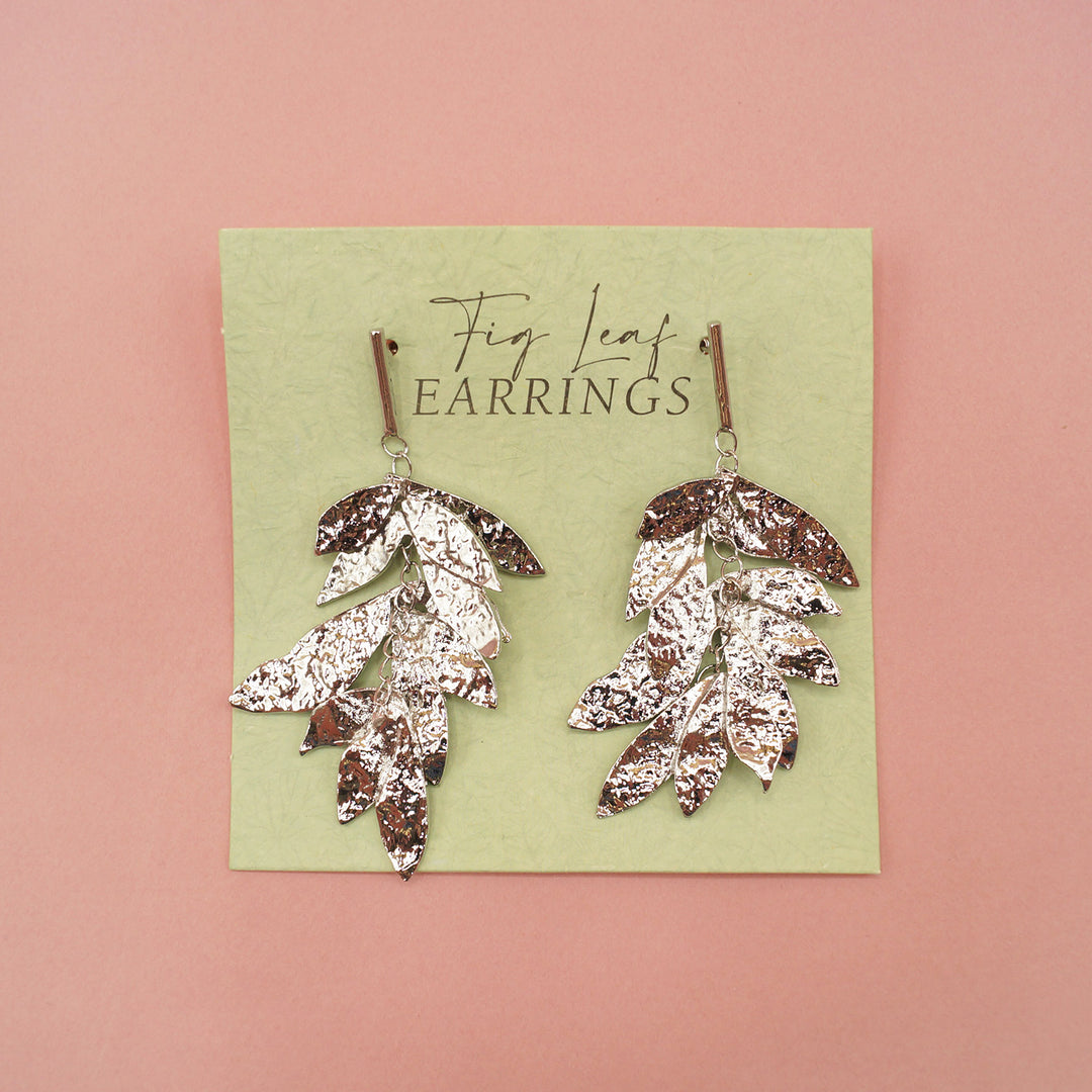 A close up of the silver dangle fig leaf earrings on a green earring card. On a pink background.