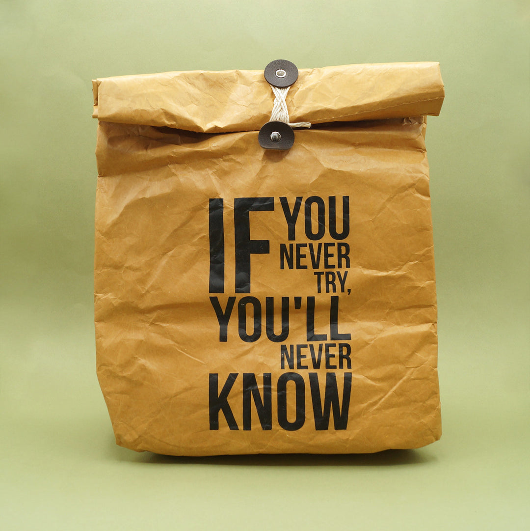 A paper reminiscent material to look like a paper lunch bag but is printed and lined. String closure. Outside printed with the words "If you never try, you'll never know."