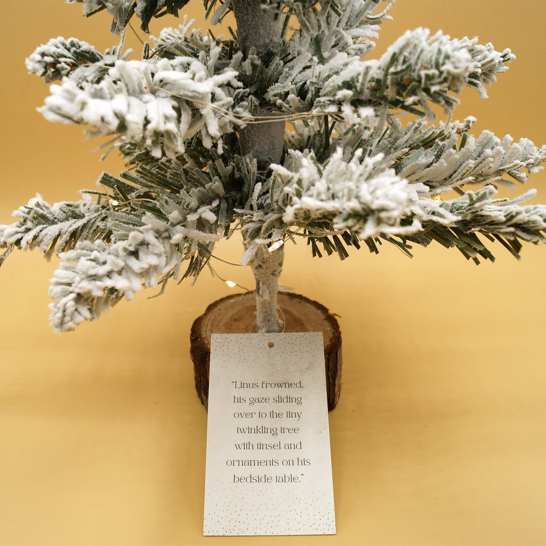 A close up of the tag attached to each miniature Christmas tree that reads "Linus frowned, his gaze sliding over to the tiny twinkling tree with tinsel and ornaments on his bedside table.
