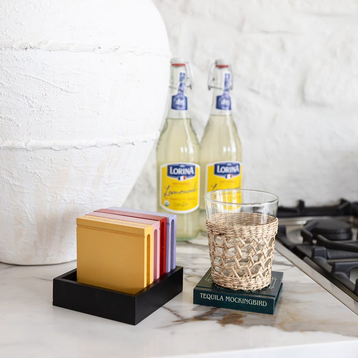 A glass sits on a wooden, book-shaped coaster featuring "Tequila Mockingbird" on the spine. The other four coasters stand ready in their stand to the left of the glass.