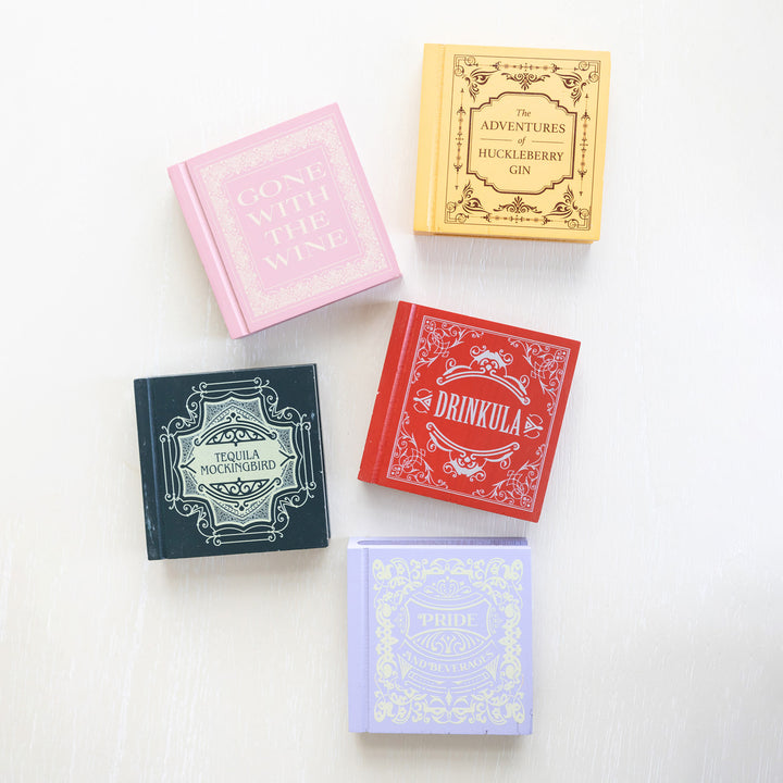 A set of five drink coasters featuring drink puns from classic novels including "Drinkula", "Tequila Mockingbird", "Gone with the Wine", "The Adventures of Huckleberry Gin", and "Pride and Beverages" in various colors.