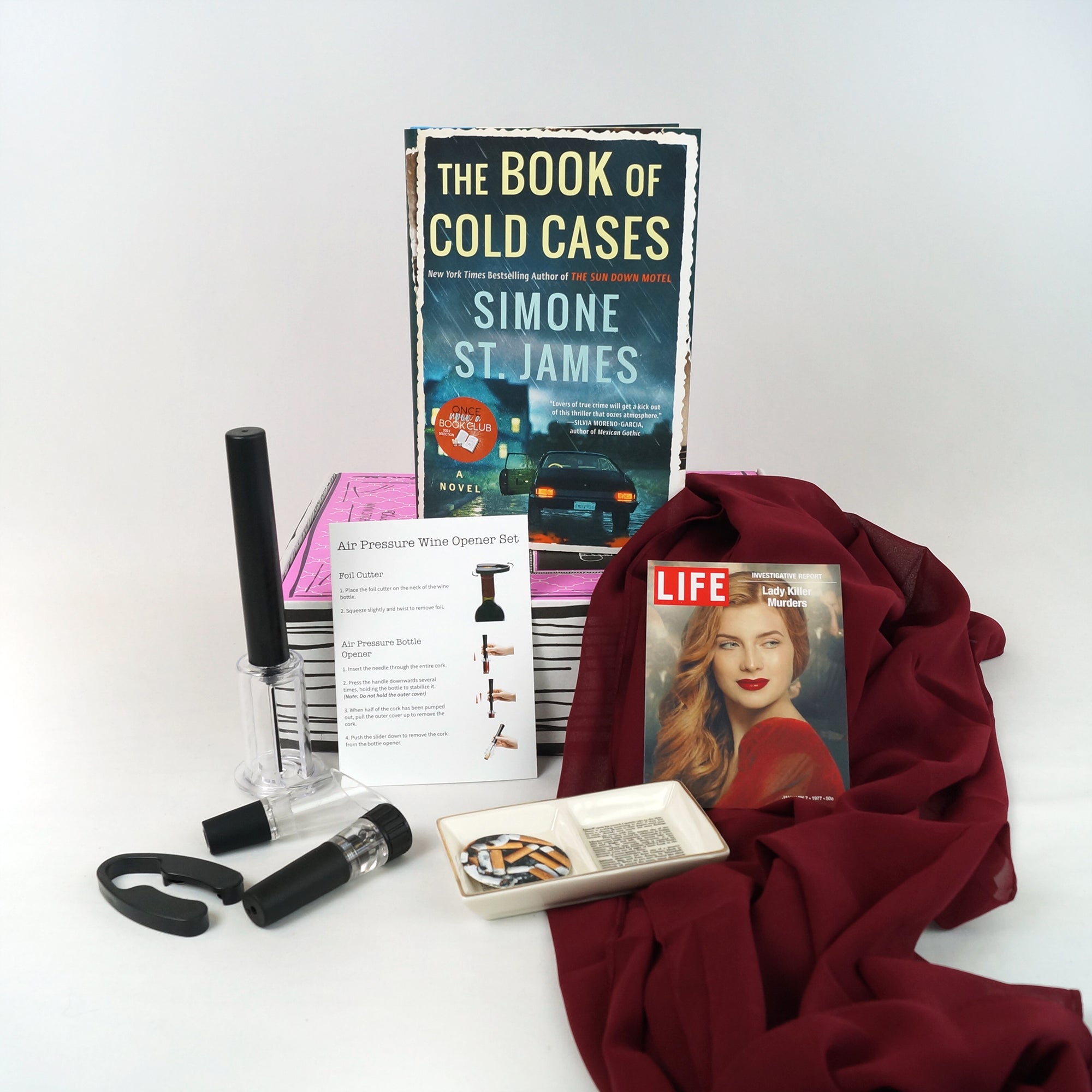 A hardcover edition of The Book of Cold Cases by Simone St. James sits on a pink box surrounded by the unwrapped gifts from the box including a wine tool kit, a book-shaped trinket dish, sheer red scarf, and Life Magazine recreation.