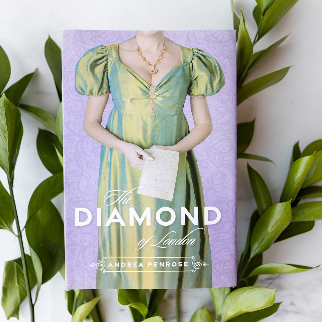 A close up at the hardcover exclusive edition of The Diamond of London by Andrea Penrose. Dust jacket designed by Once Upon a Book Club. The dust jacket features a white woman in a long, period-appropriate gown clutching a letter in her hand.