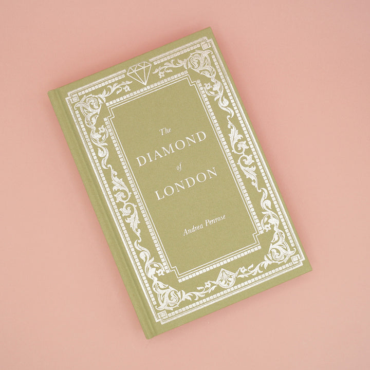 A close up of the Once Upon a Book Club exclusive edition of The Diamond of London by Andrea Penrose fabric hard case. The case is light green and features silver foiling in a classic style. A diamond gemstone is designed into the pattern at the top of the book.