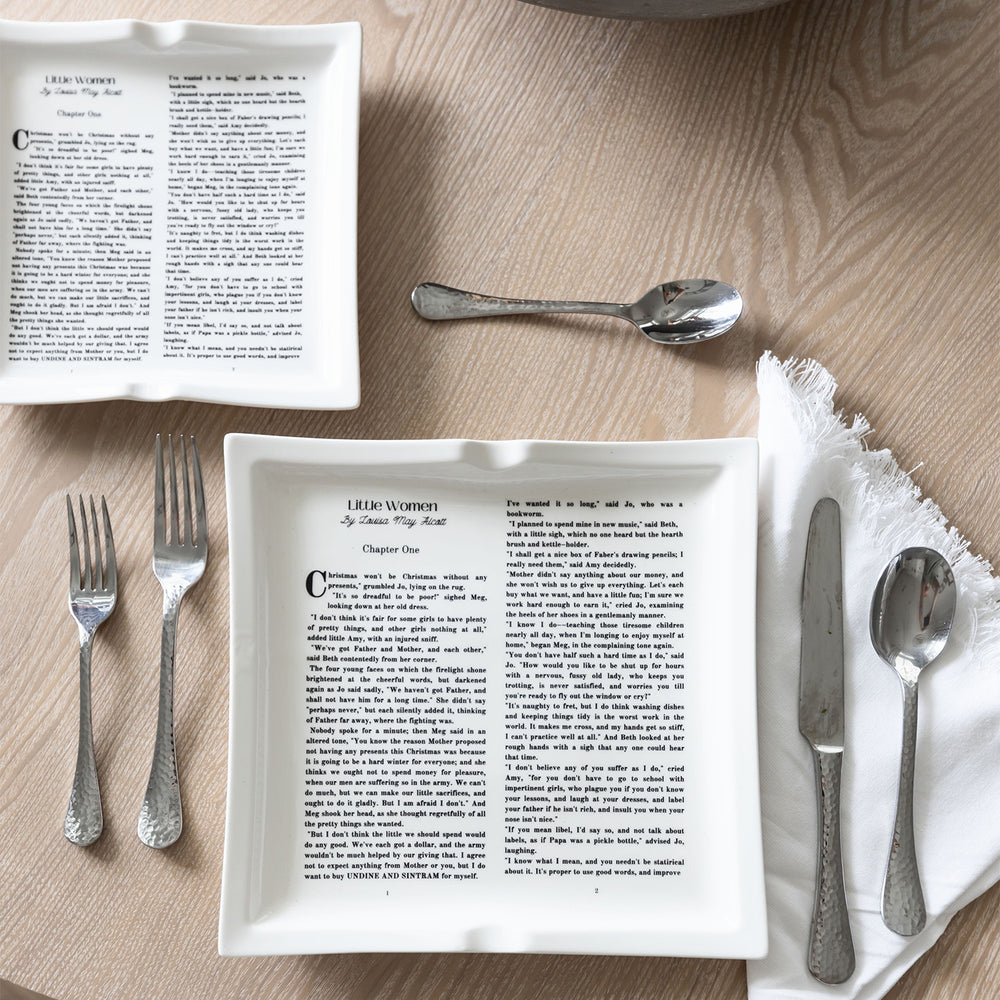Dinner plates with Little Women text, paired with silver utensils and a white napkin on a table.