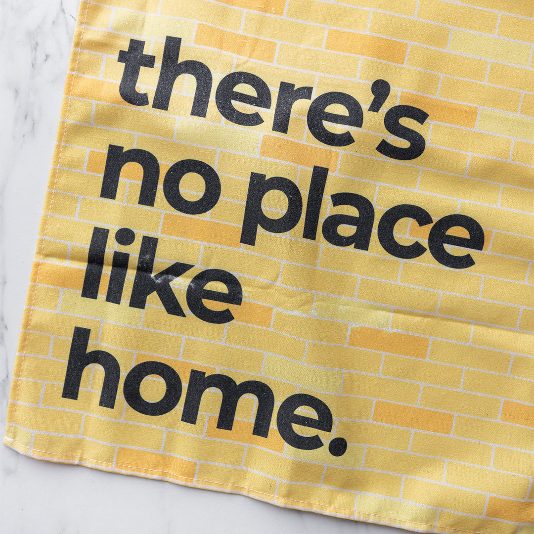 Yellow tea towel with black text "there's no place like home" on a brick-patterned background.