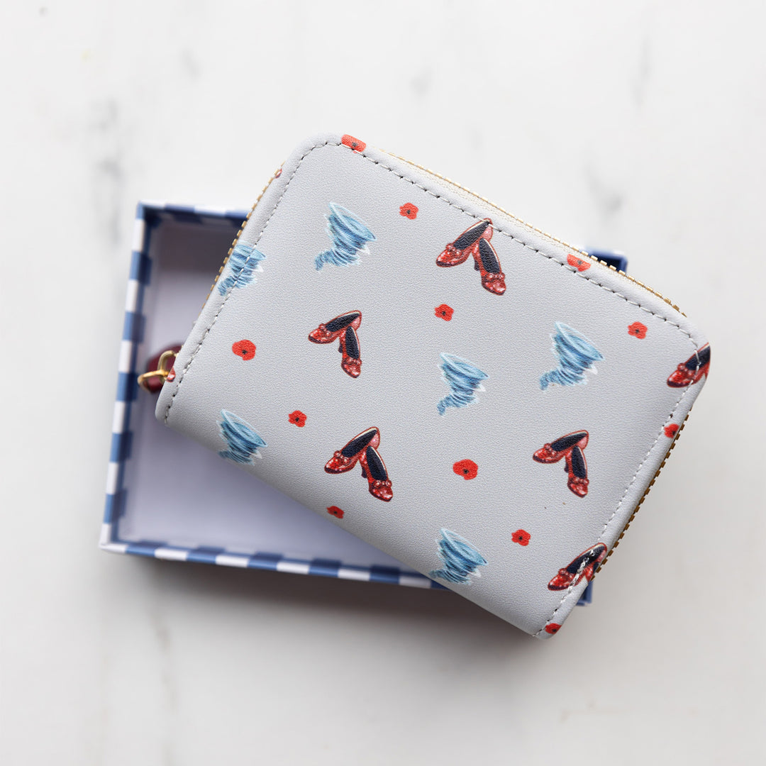 A grey wallet zipped closed. Showcases small images printed on the material of ruby red high heels, tornadoes, and poppies.