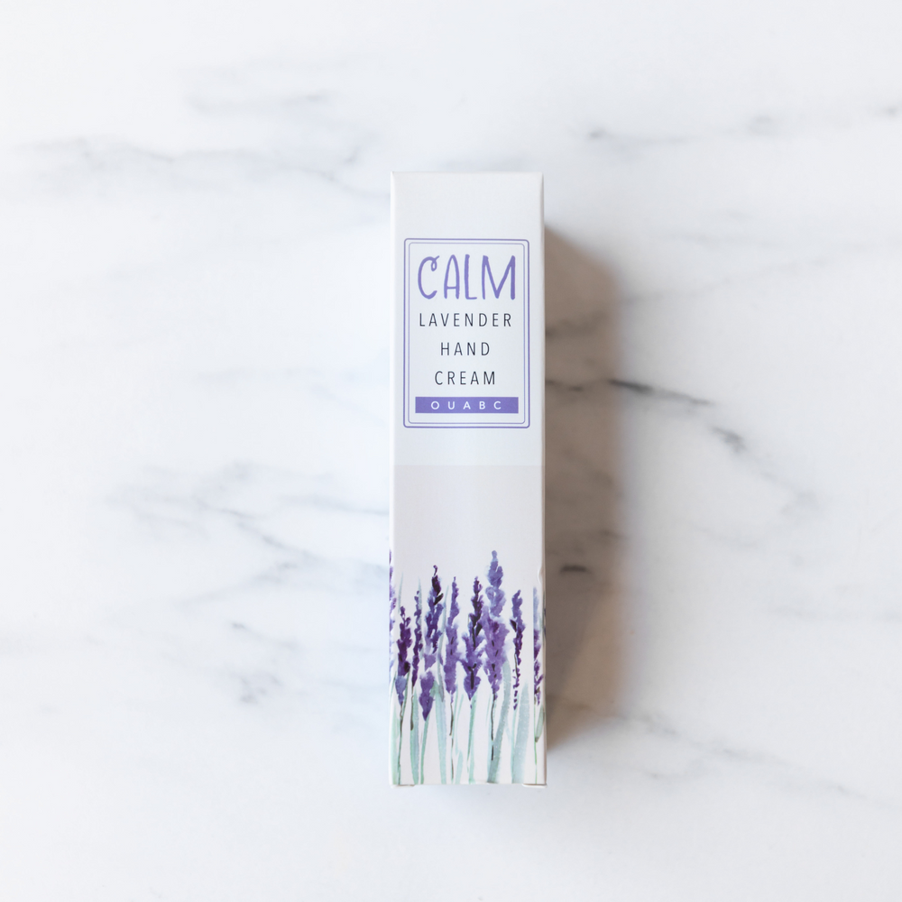 A box patterned with lavender stalks sits on a white marble background. The box reads "Calm Lavender Hand Cream. OUABC"