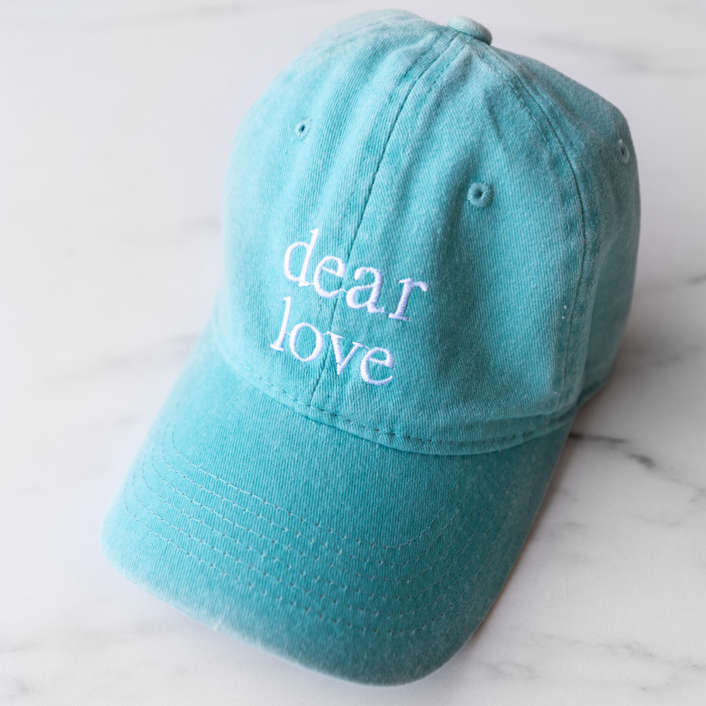 A light blue baseball hat featuring the words "Dear Love" in white embroidery thread.
