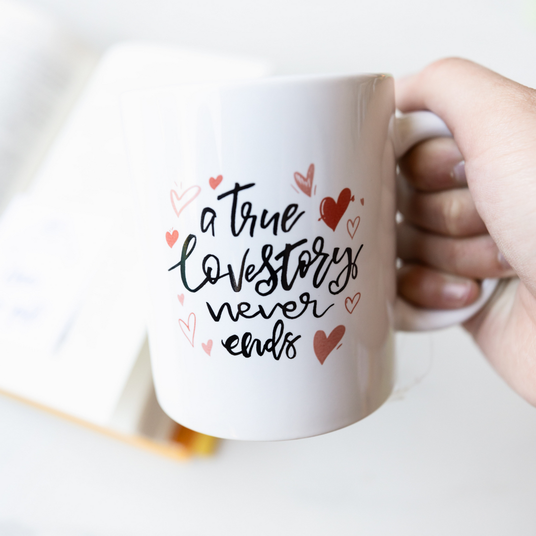 A white hand holds the handle of a white mug printed with the words "a true love story never ends" surrounded by pink and red hearts.