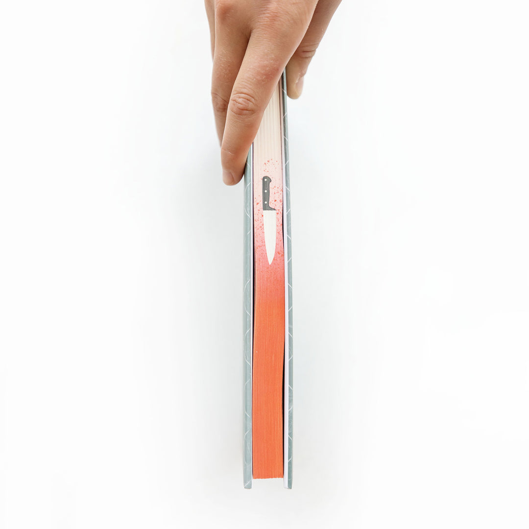 a white hand holds a hardcover book showing the page edges - red ombre with a kitchen knife