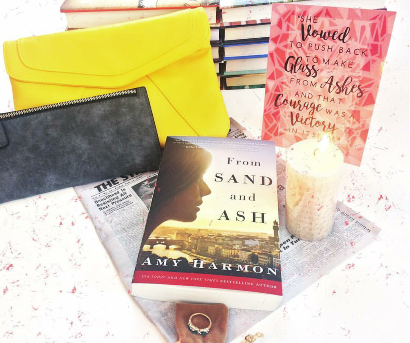 A paperback copy of From Sand and Ash by Amy Harmon sits on top of a set of unwrapped gifts inspired by the novel. These include a grey zippered wallet, a yellow crossbody purse, a candle printed with music notes, a ring, and a newspaper article.