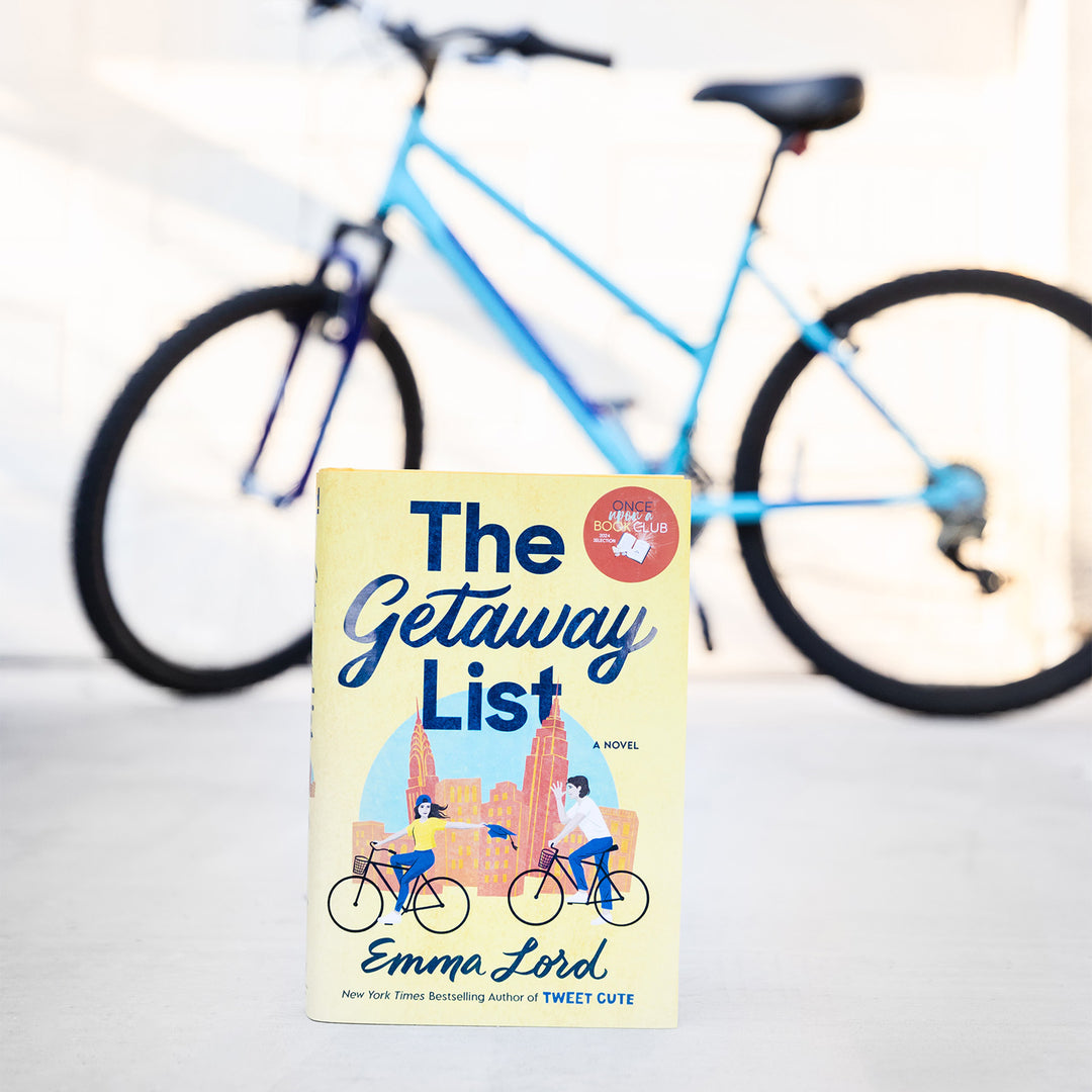A hardcover copy of The Getaway List by Emma Lord sits outside in front of a blue bike.