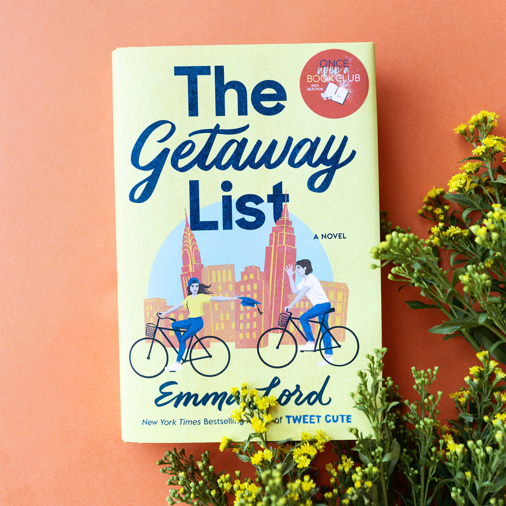 A hardcover copy of The Getaway List by Emma Lord sits on a orange background with yellow flowers in the bottom right corner.