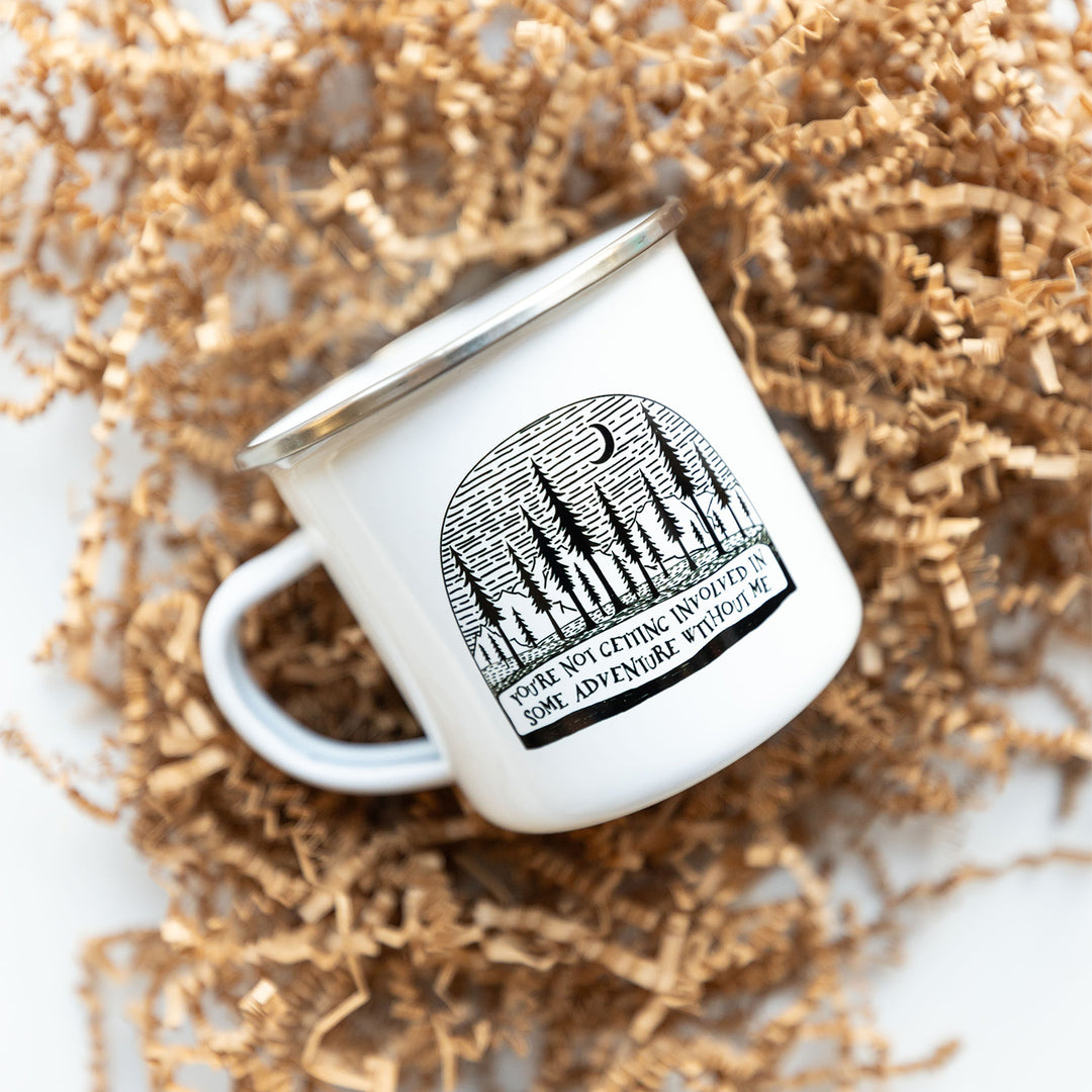 A white enamel mug sits on a bed of crinkle paper. A tree scene with the moon visible is printed on the mug. The words along the bottom of the mug says "You're not getting involved in some adventure without me"