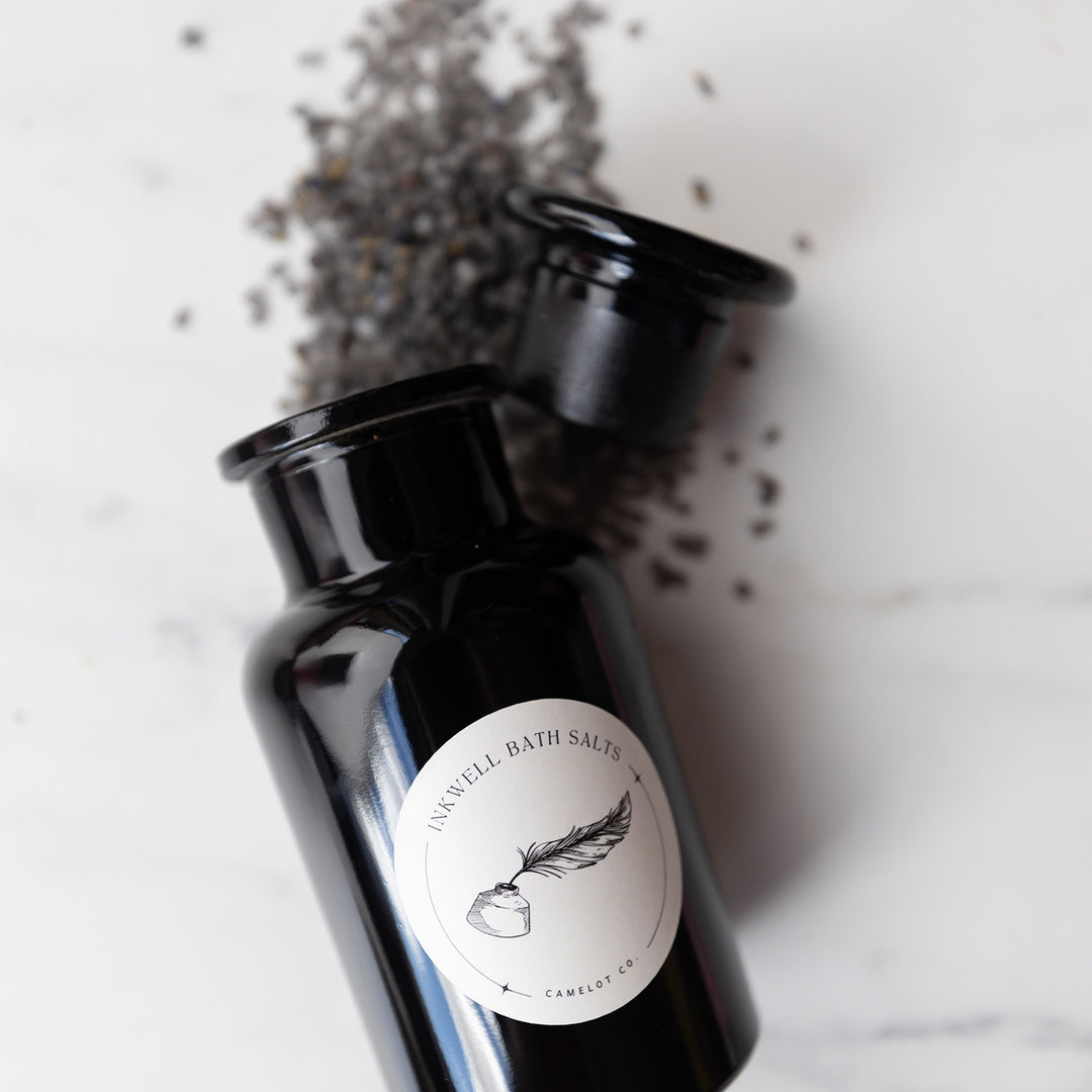 A close up of the black jar showcasing the label "Inkwell Bath Salts, Camelot Co" and the image of a quill in ink in the middle of it. The jar is open and bath salts are spilling from the top.