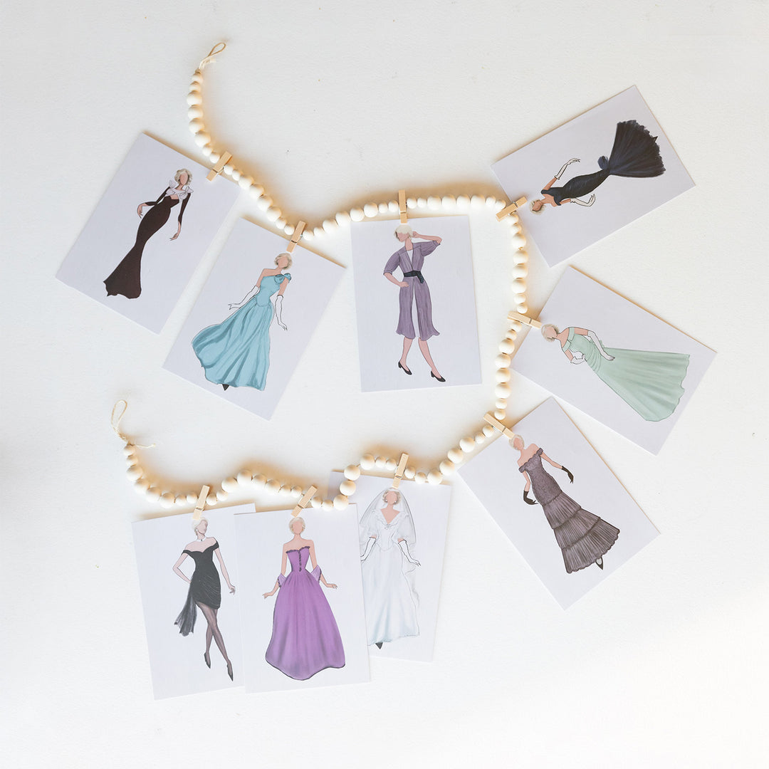 A string of white beads with nine clothespins on it evenly spaced out. Each clothespin holds an art print of a woman wearing a dress