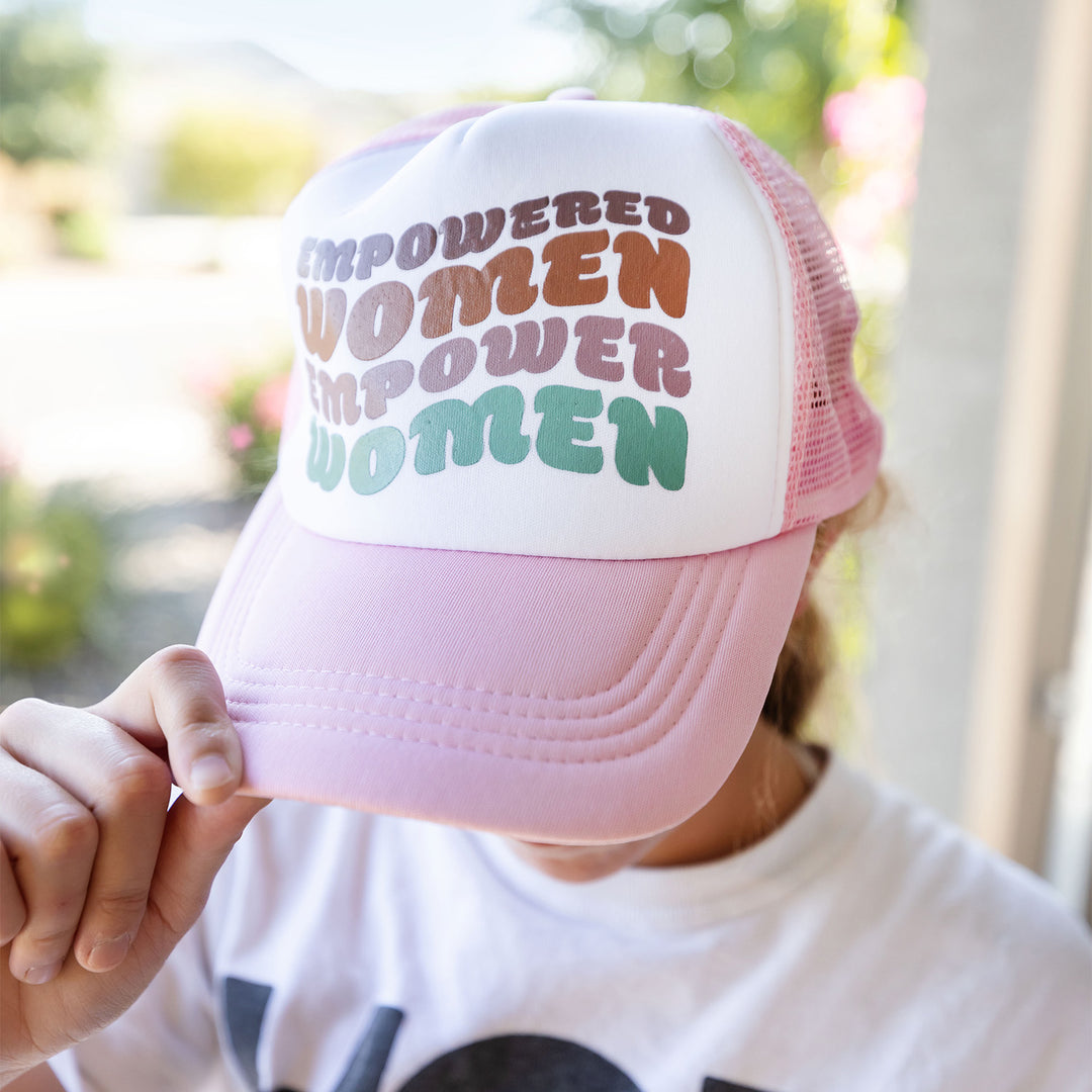a woman wearing a pink snapback trucker hat that says "empowered women empower women" on the front, holding the brim of the hat